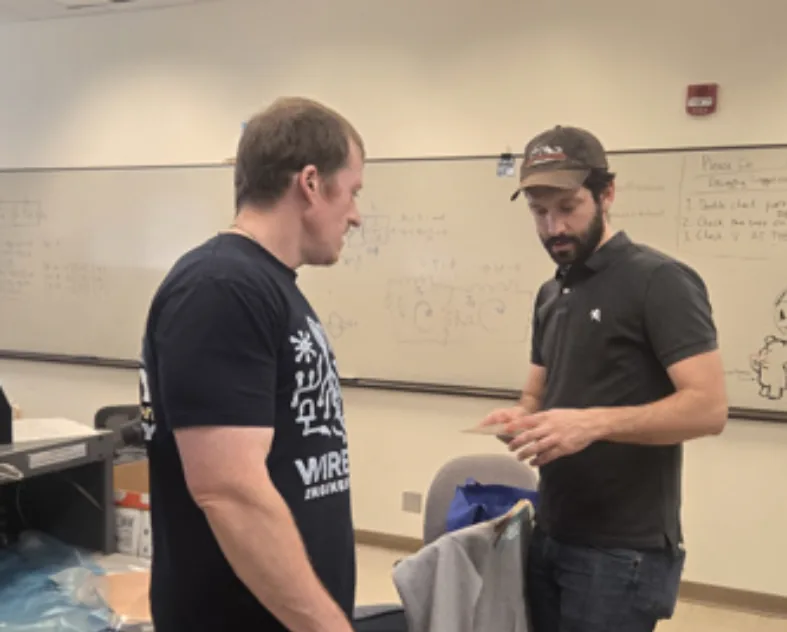 Two members reviewing a board in front of a whiteboard covered in circuit sketches